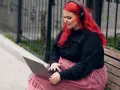 Młoda babista z ognistymi włosami i słuchawkami na uszach pracuje na laptopie, siedząc na ławce w parku.