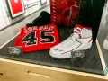 Białe buty sportowe z czerwonymi akcentami, podpisane przez Michaela Jordana, obok koszulki z numerem 45. To pamiątki, które pokazują, jaki michael jordan rozmiar buta nosił.