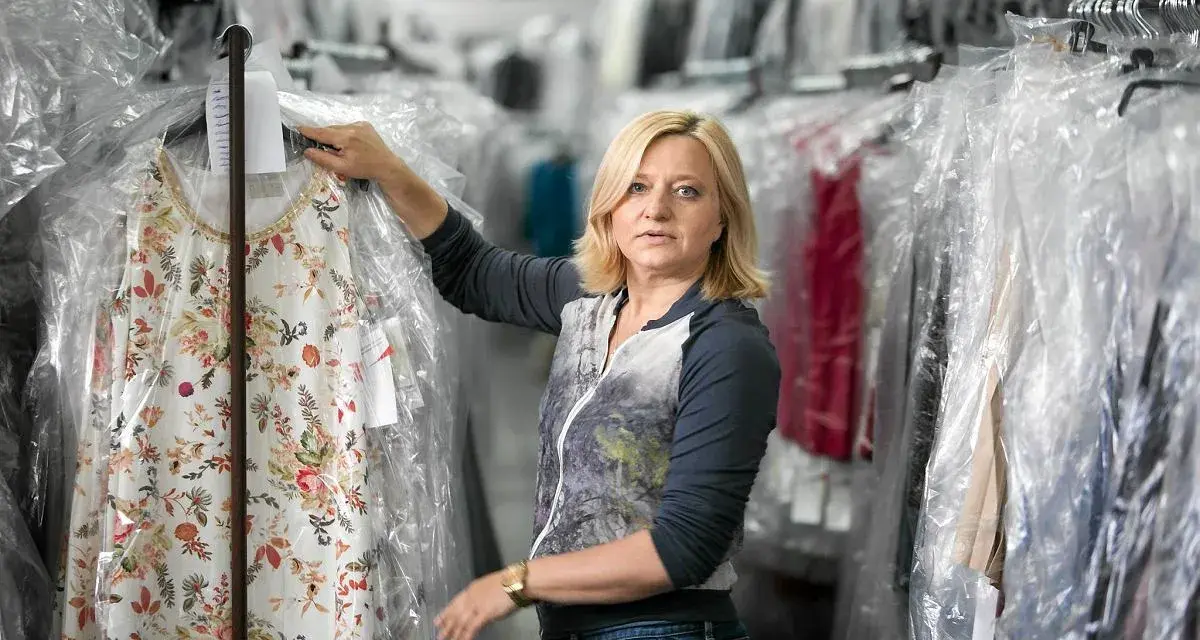 Blondynka w magazynie pralni, tu woman co to za firma? Wieszak z kwiecistą sukienką w folii.