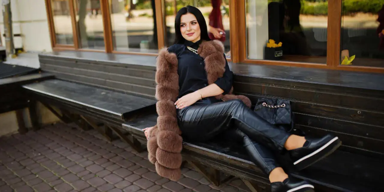 Elegancka kobieta w futrzanej kamizelce i skórzanych spodniach, zastanawia się, jakie buty do skorzanych spodni będą najlepsze. Wybrała czarne, platformowe mokasyny.