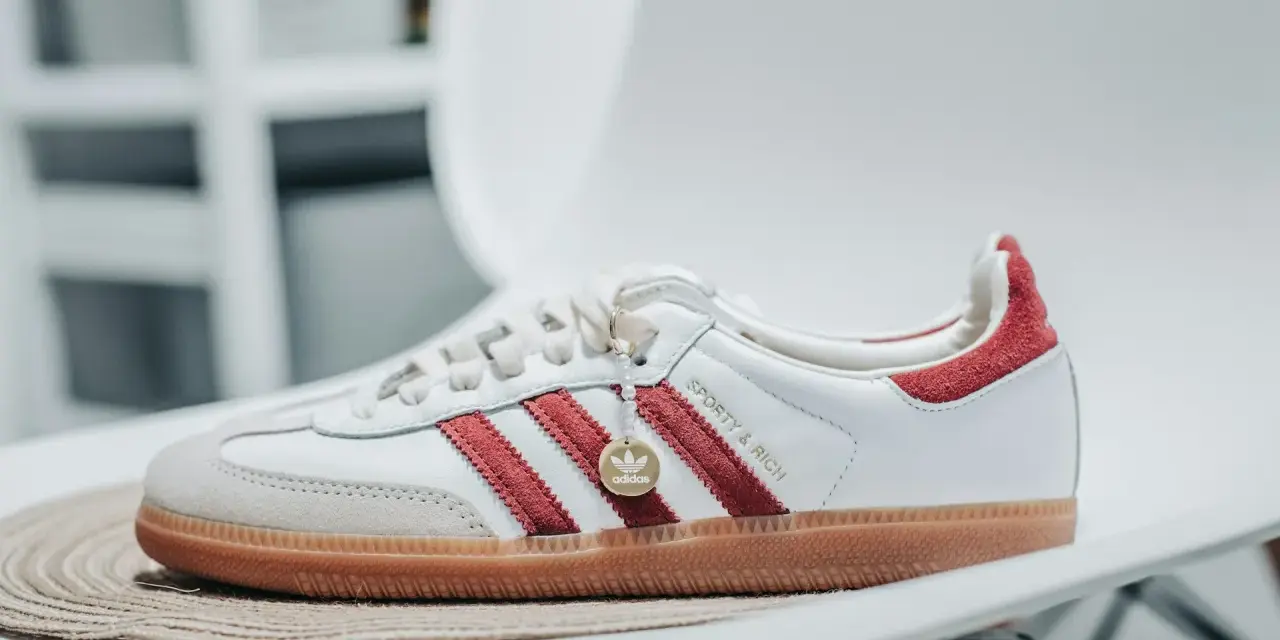 Białe sneakersy Adidas z czerwonymi paskami i zamszowym noskiem. Złoty medalik z logo Adidas i napis "Sporty & Rich" sugerują idealną rozmiarówkę Adidasa.