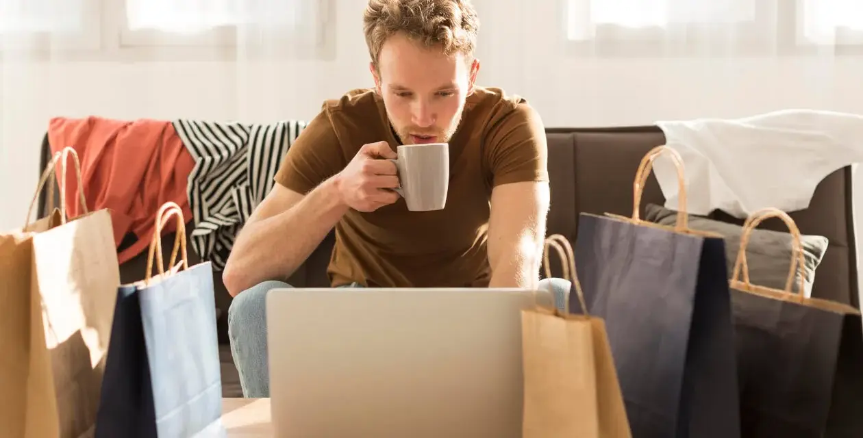 Mężczyzna popija kawę, przeglądając laptopa. Wokół niego leżą torby zakupowe i ubrania, co sugeruje, że szuka odpowiedzi, czy na Zalando Lounge są oryginalne rzeczy.