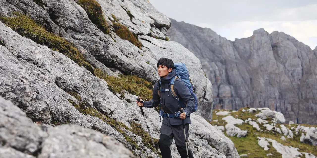 Mężczyzna w kurtce i spodniach trekkingowych z plecakiem wspina się po skalistym zboczu. Idealny przykład, jak się ubrać w góry wiosną.