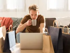 Mężczyzna popija kawę, przeglądając laptopa. Wokół niego leżą torby zakupowe i ubrania, co sugeruje, że szuka odpowiedzi, czy na Zalando Lounge są oryginalne rzeczy.