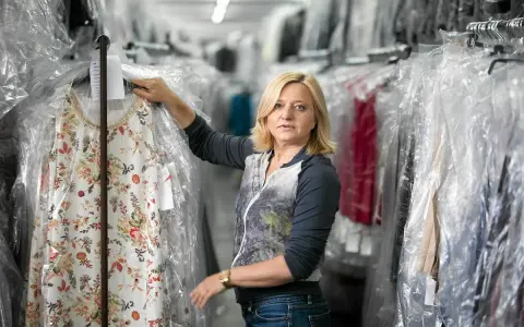Blondynka w magazynie pralni, tu woman co to za firma? Wieszak z kwiecistą sukienką w folii.