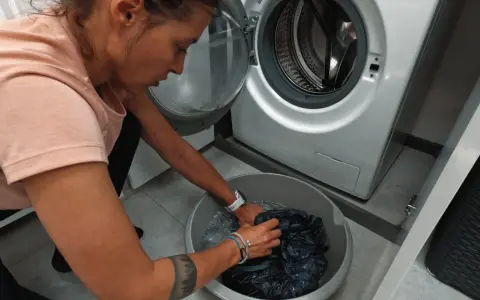 Kobieta w różowej koszulce pierze ciemne ubrania w misce obok pralki. Może to być część rutyny sprzątania w domu w Kilmanock.