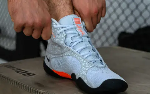 Ręce zapinają jasnoniebieskie buty sportowe z pomarańczowymi akcentami, przypominające styl Batistini.