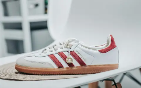 Białe sneakersy Adidas z czerwonymi paskami i zamszowym noskiem. Złoty medalik z logo Adidas i napis "Sporty & Rich" sugerują idealną rozmiarówkę Adidasa.