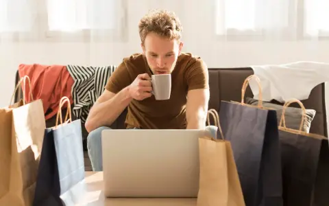 Mężczyzna popija kawę, przeglądając laptopa. Wokół niego leżą torby zakupowe i ubrania, co sugeruje, że szuka odpowiedzi, czy na Zalando Lounge są oryginalne rzeczy.