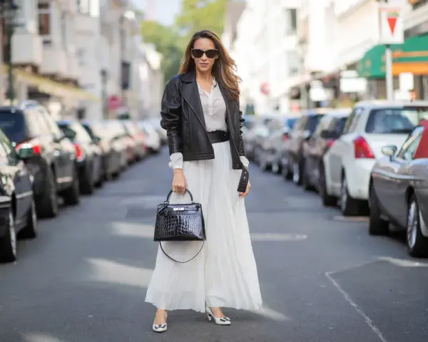 Elegancka kobieta w białej, plisowanej spódnicy i czarnej ramonesce. Idealne buty do plisowanej spódnicy to te, które widzisz: białe czółenka z czarną kokardką.