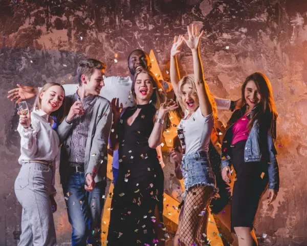 Grupa młodych ludzi świętuje, sypiąc konfetti. Inspiracja, jak się ubrać na imprezę do klubu: sukienka, jeansy, topy, marynarka.