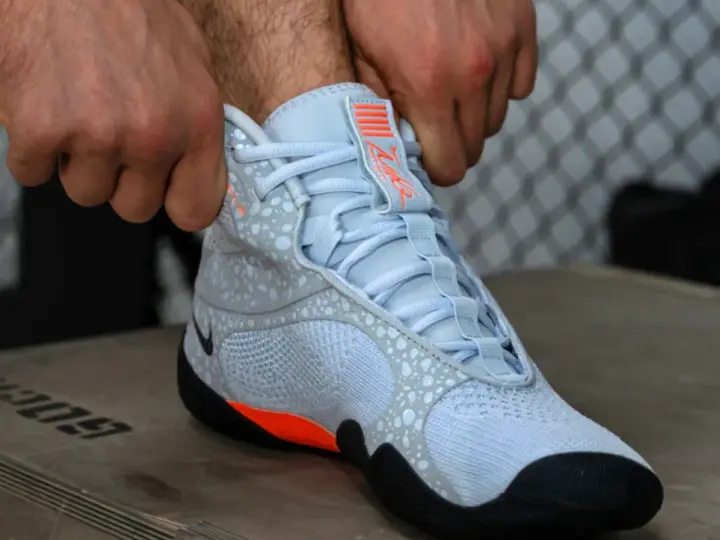 Ręce zapinają jasnoniebieskie buty sportowe z pomarańczowymi akcentami, przypominające styl Batistini.