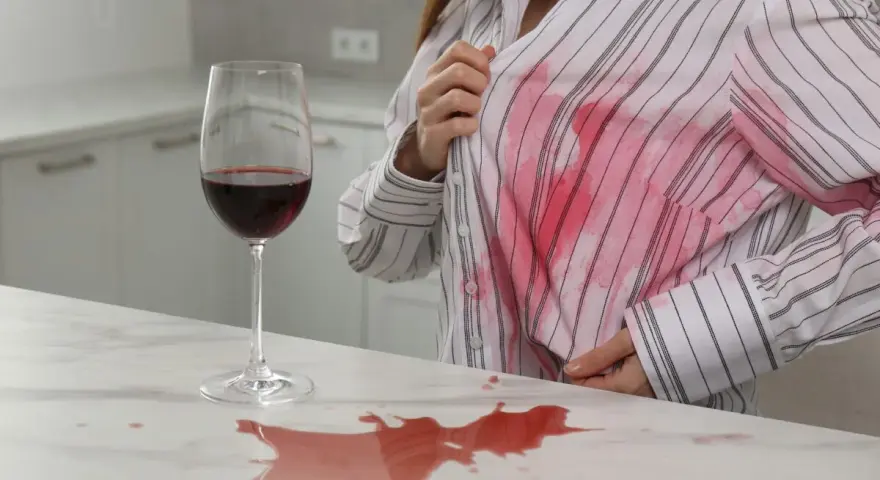 Kobieta z przerażeniem patrzy na plamę z czerwonego wina na swojej białej koszuli. Dowiedz się, jak usunąć plamę z wina.