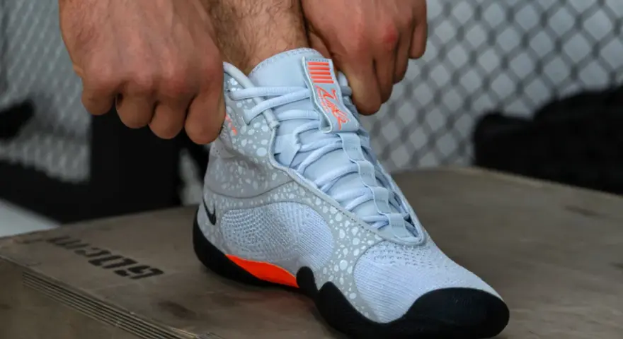 Ręce zapinają jasnoniebieskie buty sportowe z pomarańczowymi akcentami, przypominające styl Batistini.