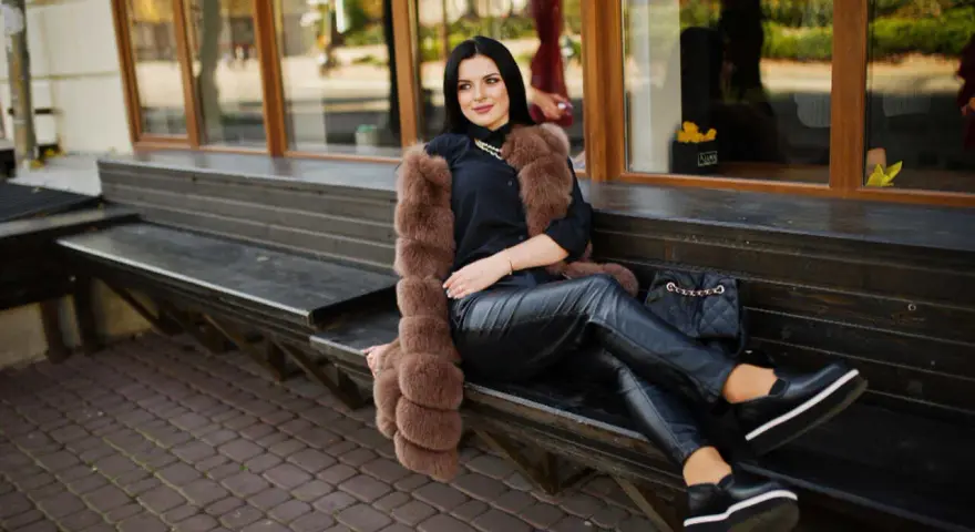 Elegancka kobieta w futrzanej kamizelce i skórzanych spodniach, zastanawia się, jakie buty do skorzanych spodni będą najlepsze. Wybrała czarne, platformowe mokasyny.