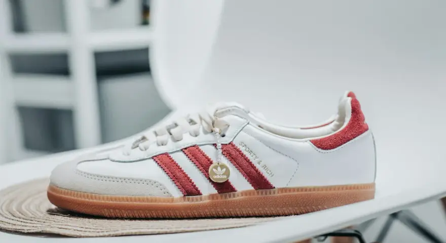 Białe sneakersy Adidas z czerwonymi paskami i zamszowym noskiem. Złoty medalik z logo Adidas i napis "Sporty & Rich" sugerują idealną rozmiarówkę Adidasa.