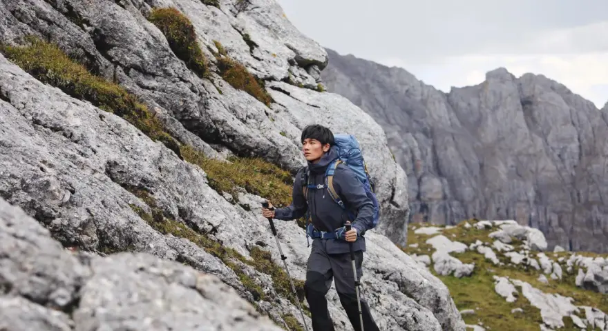 Mężczyzna w kurtce i spodniach trekkingowych z plecakiem wspina się po skalistym zboczu. Idealny przykład, jak się ubrać w góry wiosną.