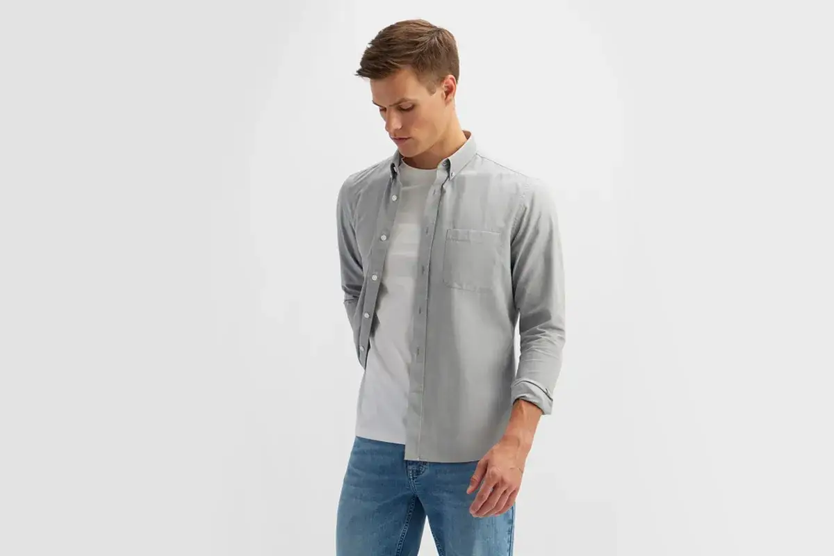 Młody mężczyzna w szarej koszuli z bawełny, pod którą ma biały t-shirt.