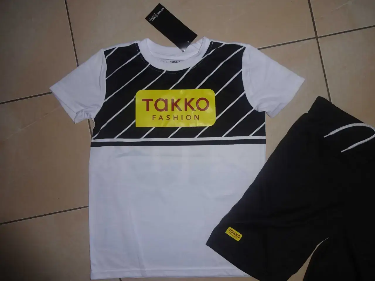 Komplet dziecięcy: biała koszulka z czarnymi pasami i żółtym logo Takko Fashion, czarne spodenki. Metka z napisem chapter young.