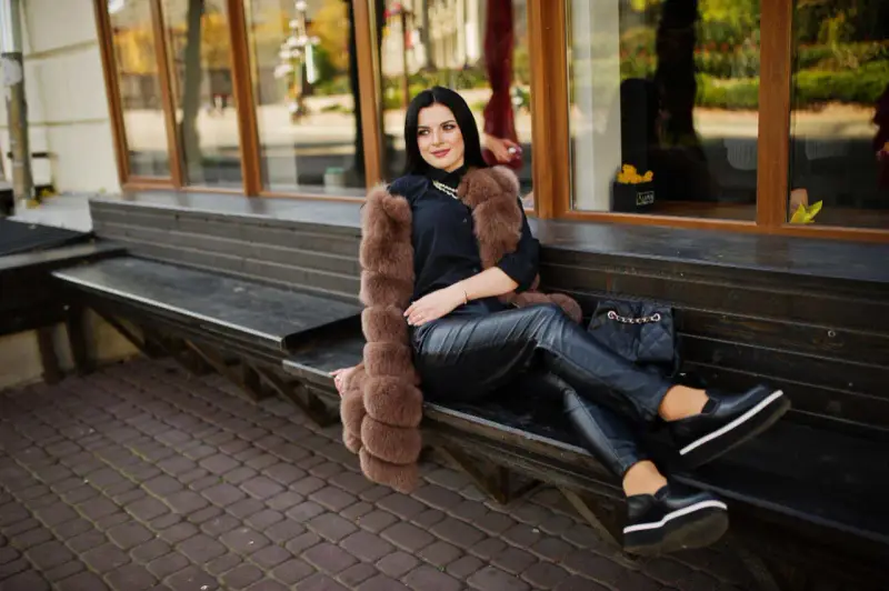 Elegancka kobieta w futrzanej kamizelce i skórzanych spodniach, zastanawia się, jakie buty do skorzanych spodni będą najlepsze. Wybrała czarne, platformowe mokasyny.
